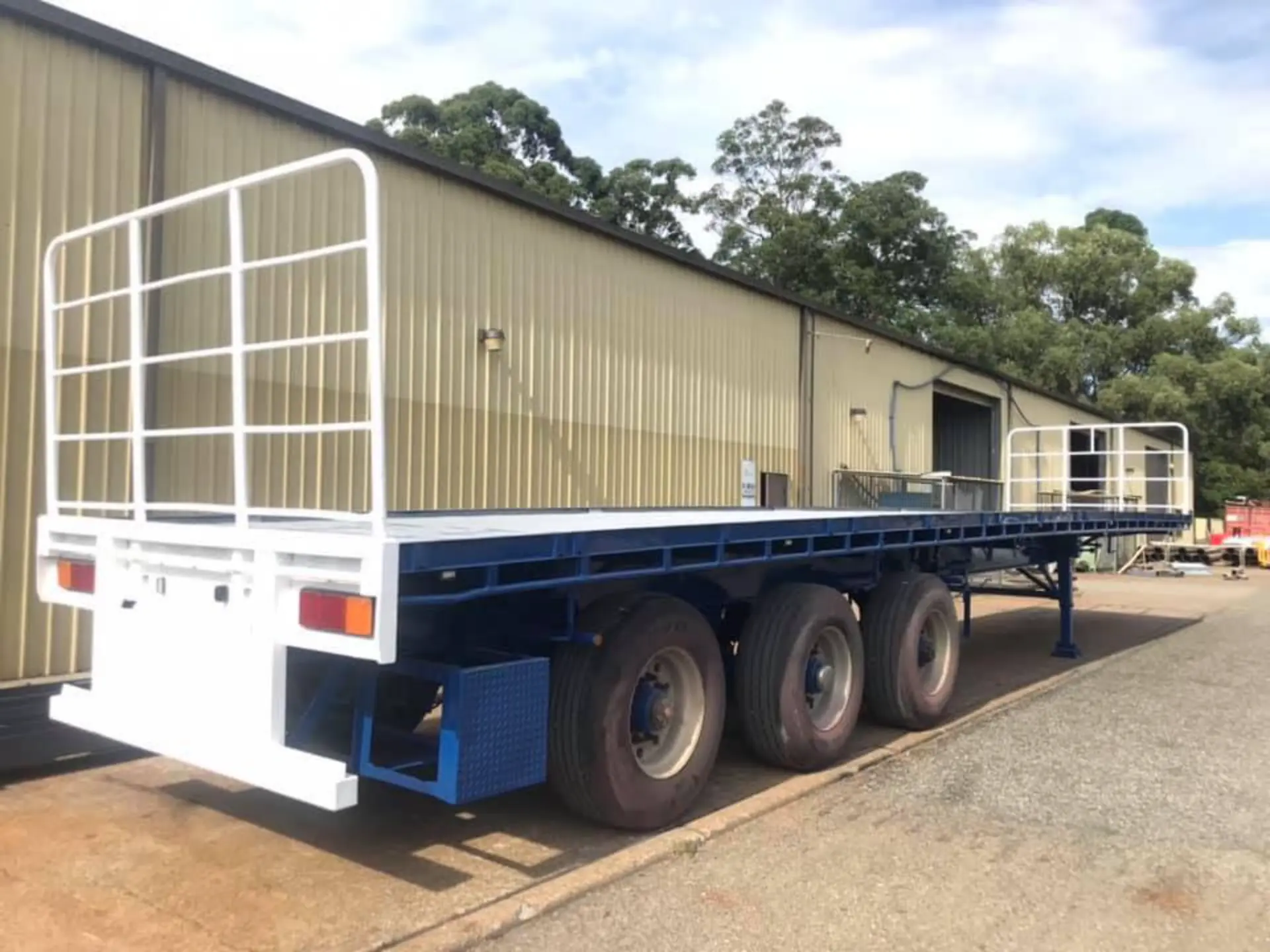 Finished flat-top trailer in fresh white and blue coating parked beside the workshop shed